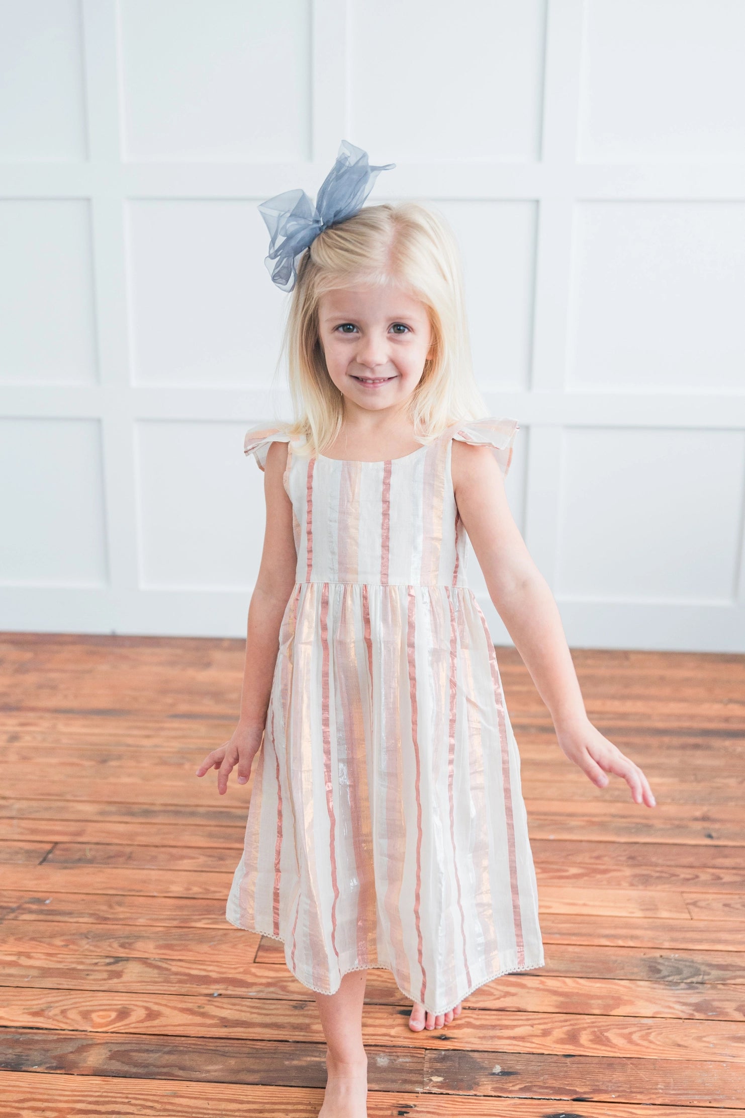Young girl in a striped dress with a blue bow standing on a wooden floor against a white wall.