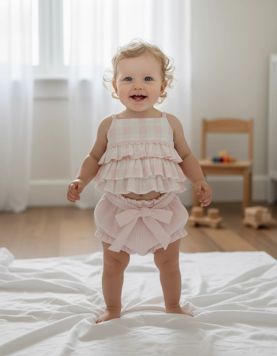 Child wearing a pink ruffled plaid top and striped shorts with a bow detail.