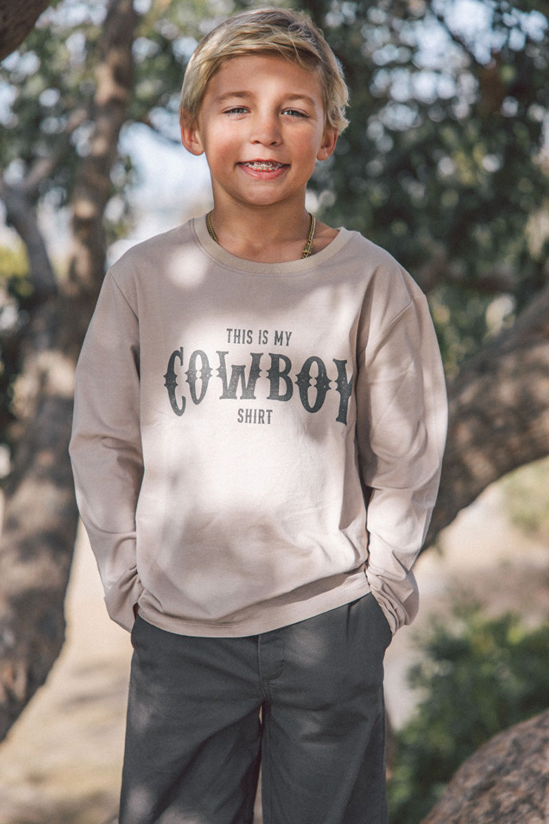 Young boy wearing a beige sweatshirt with 'This is my cowboy shirt' text, standing outdoors.