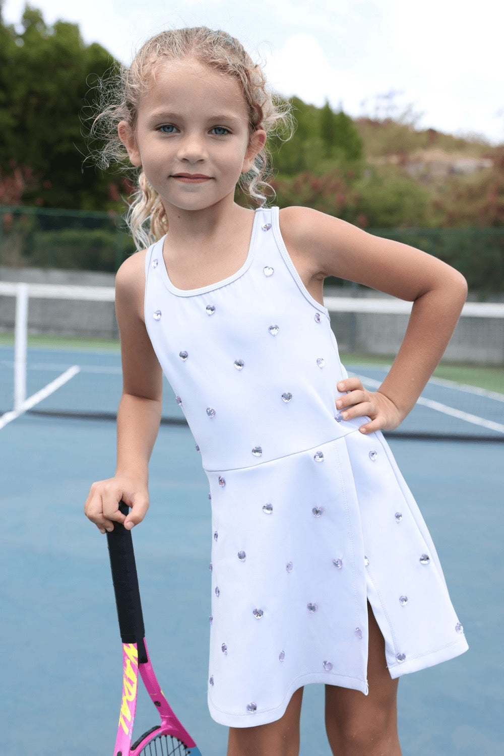 Young girl in a white dress with diamond details holding a tennis racket on a tennis court.