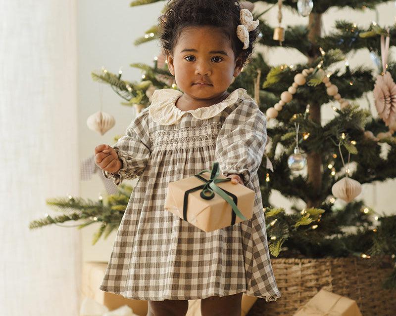 Child in a checkered dress holding a gift box in front of a decorated Christmas tree.