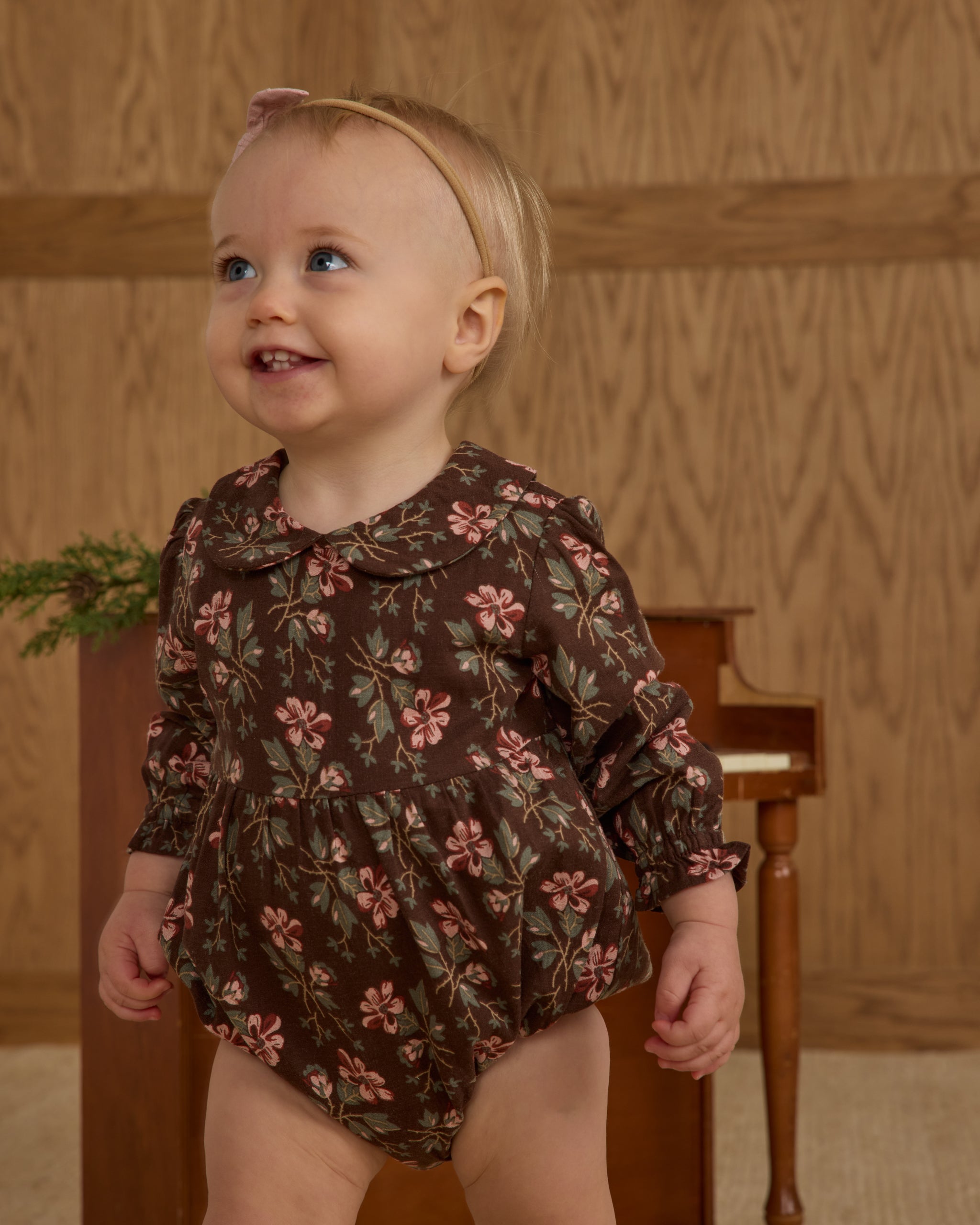 Baby wearing a floral onesie standing in front of a wooden background