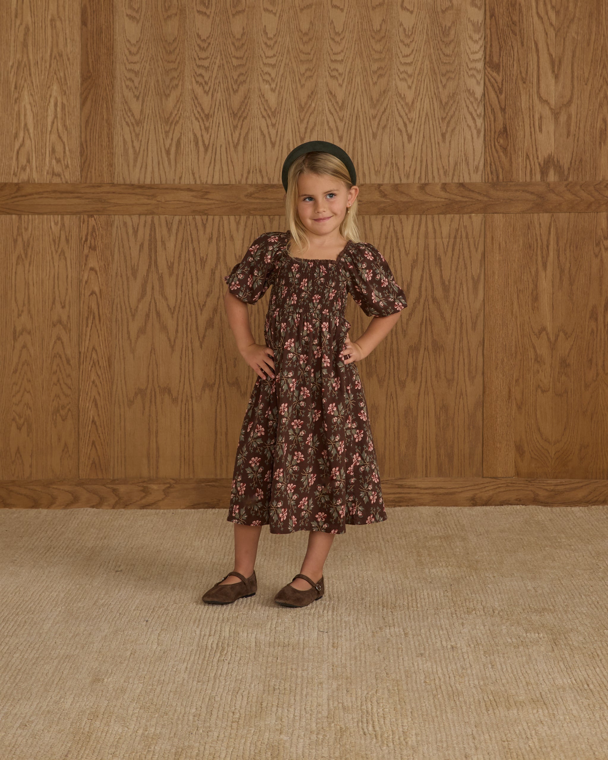 Young girl in a floral dress standing against a wooden paneled wall.