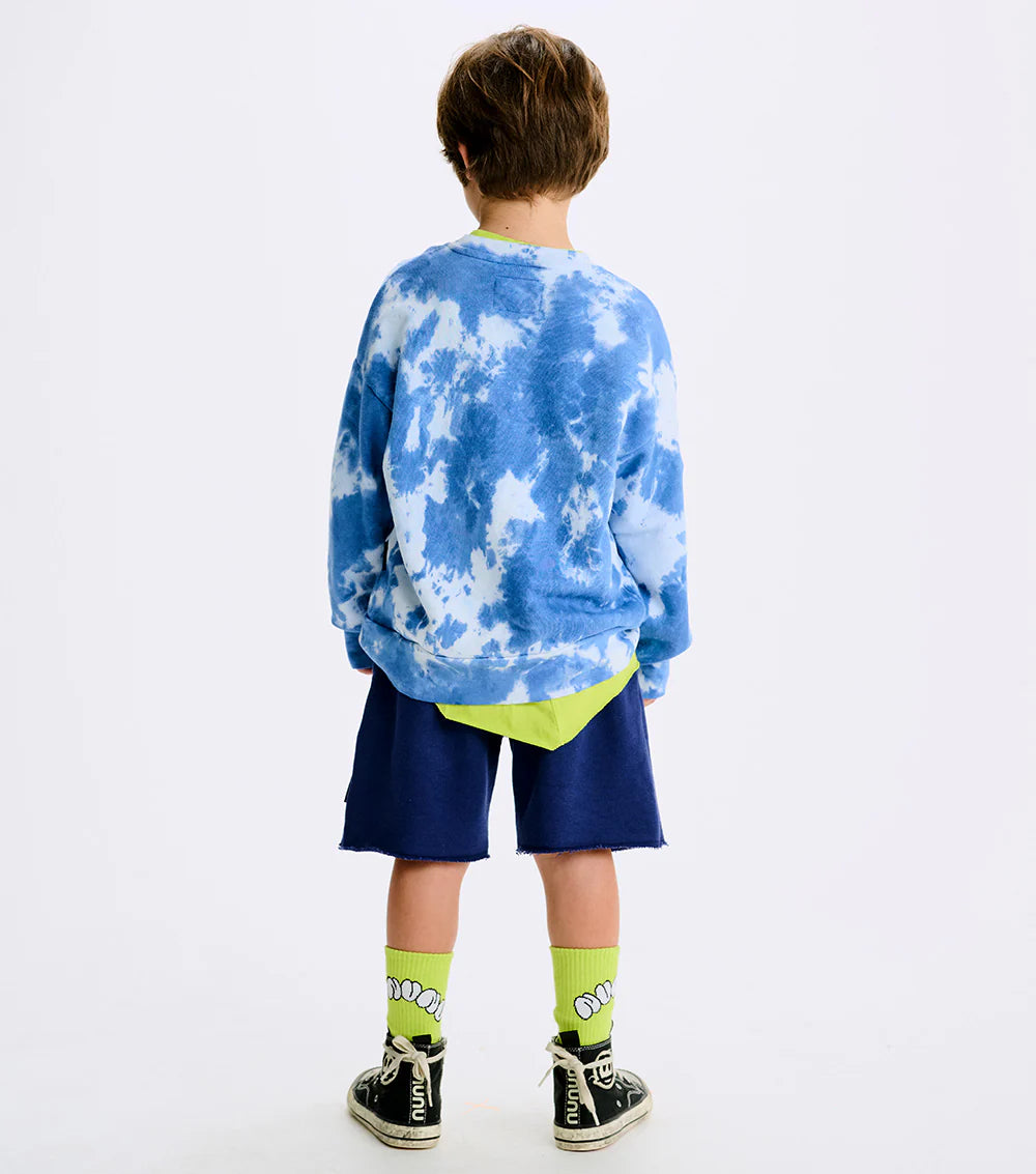 little boy standing backwards wearing a tie dye sweatshirt with blue shorts