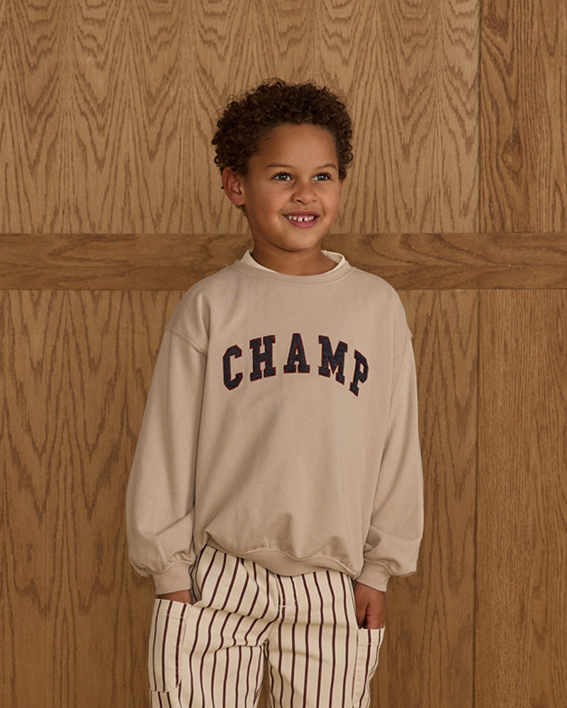 Child wearing a beige sweatshirt with 'CHAMP' text against a wooden background