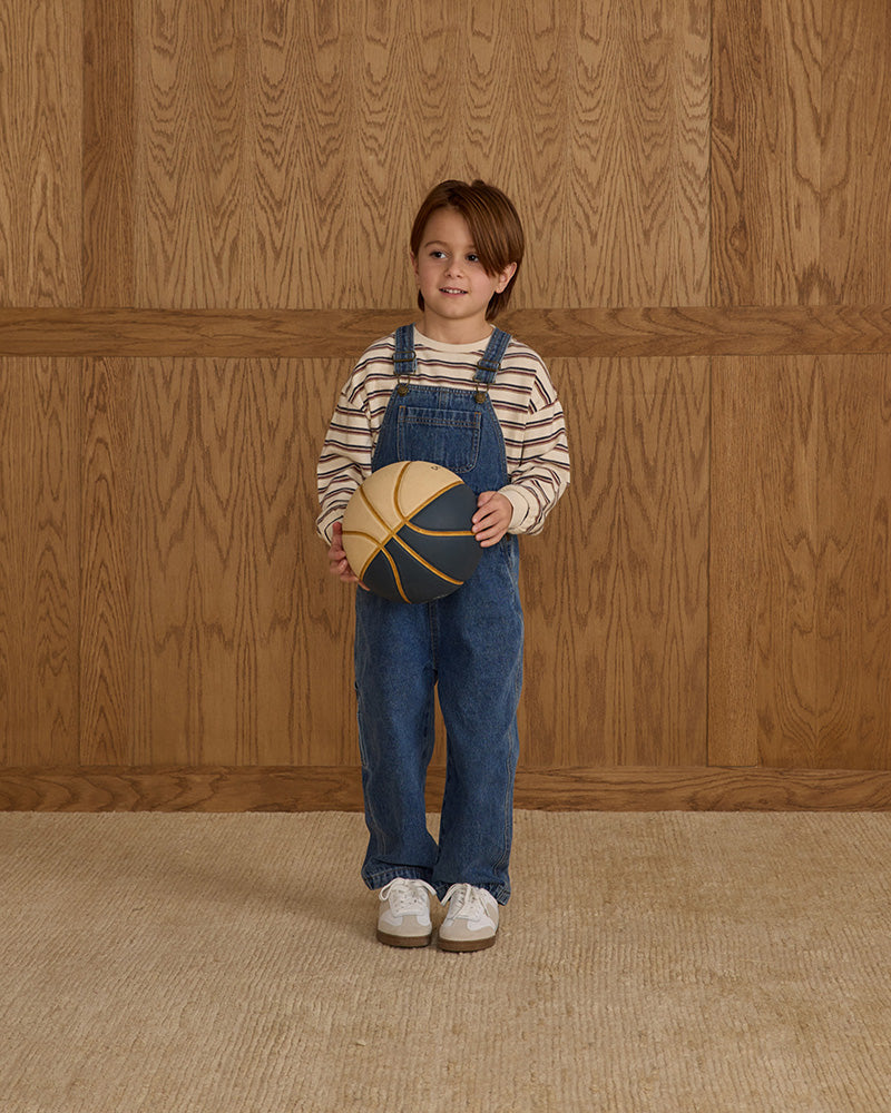Child wearing blue overalls holding a yellow and brown object against a wooden paneled wall.