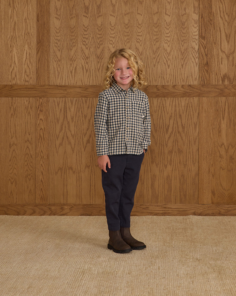 Child wearing a checkered shirt and dark pants standing against a wooden paneled wall.