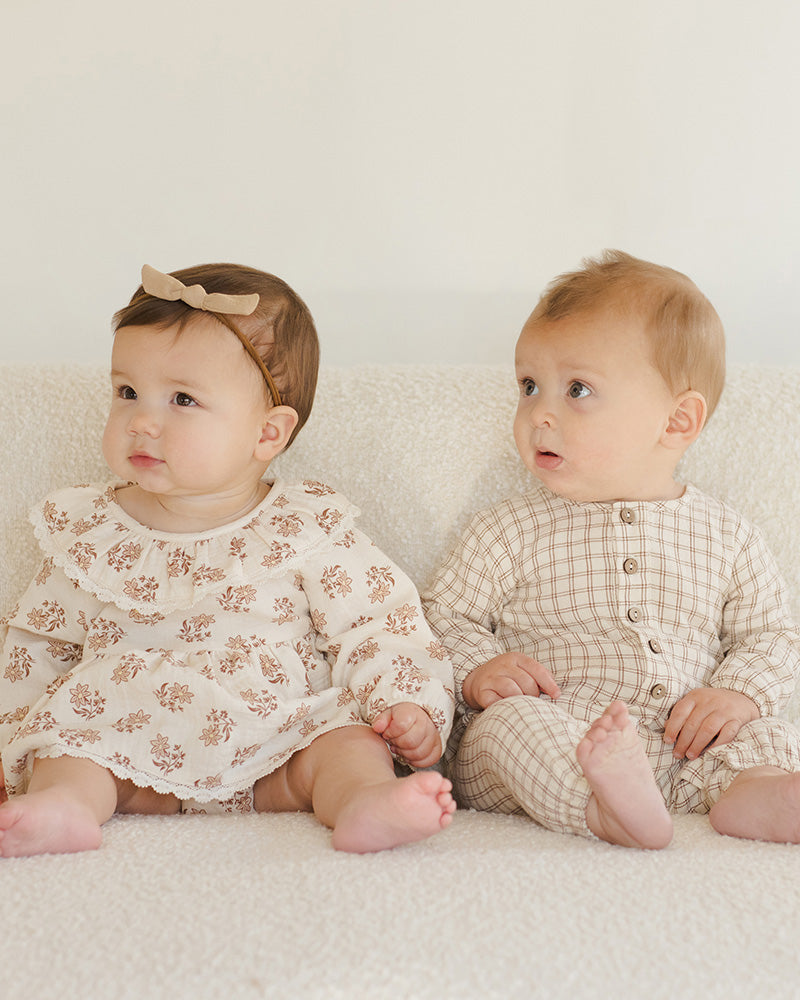 Two babies sitting on a white surface, one wearing a floral dress and the other in a checkered outfit.