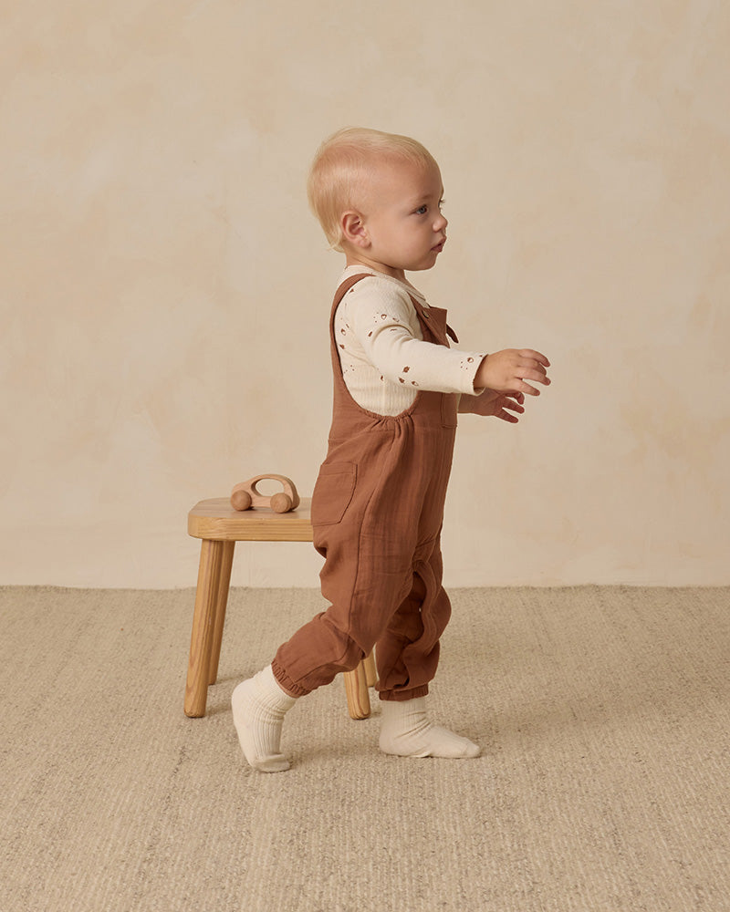 Child wearing brown overalls standing on a wooden stool against a beige background