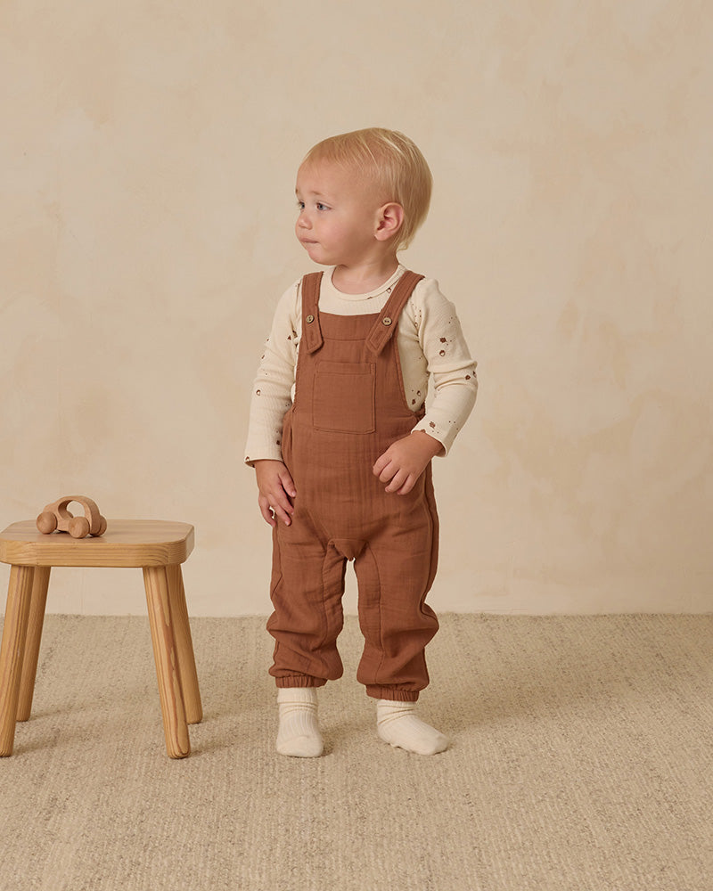 Child wearing brown overalls standing next to a wooden stool on a beige carpet.