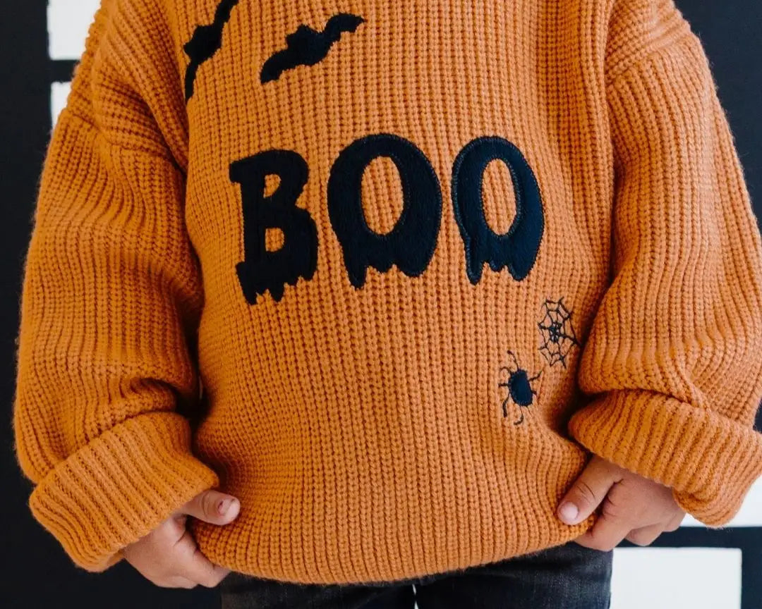Child wearing an orange sweater with 'BOO' and bat designs on a black background