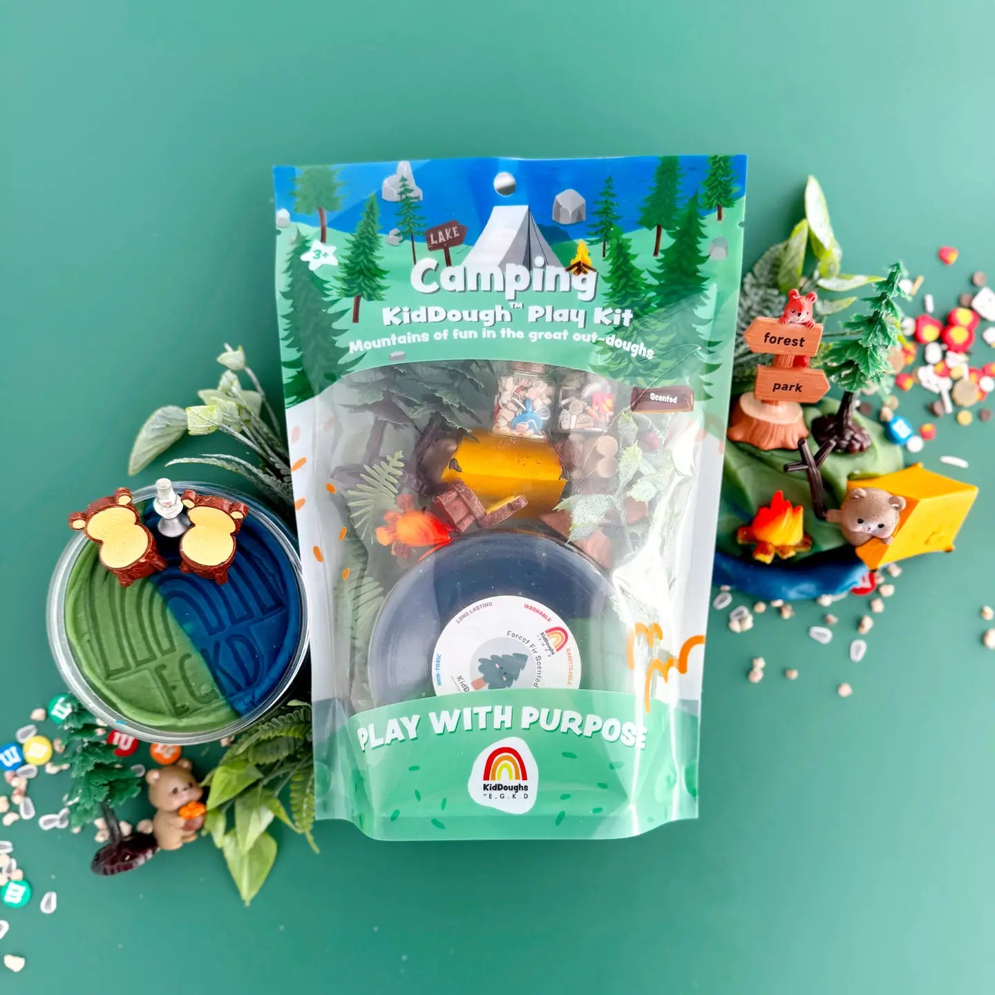 The image shows a Camping KidDough Play Kit which includes various items such as green and blue dough, a bear figurine, a tent, a fire, a stick, foliage, trees, a bear chair, a lantern, s'mores, and trail mix, all displayed on a green background.