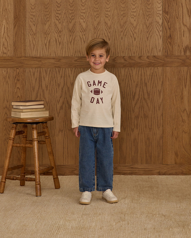 Child wearing a 'Game Day' shirt standing in front of a wooden wall with a stool and books.