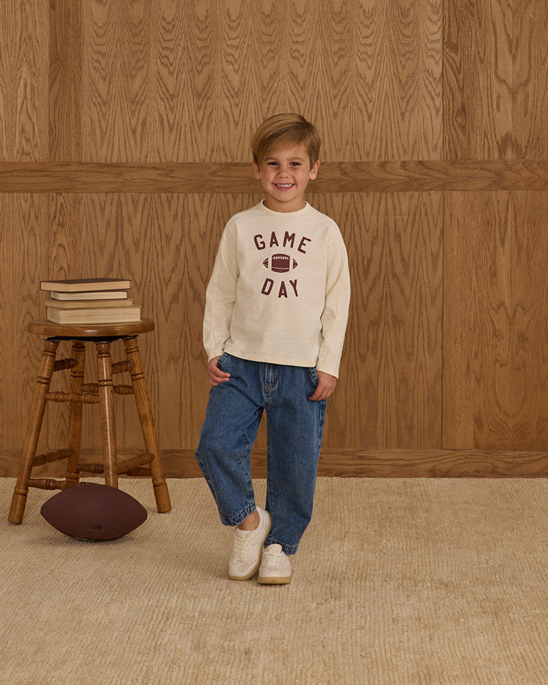 Child wearing a 'Game Day' sweatshirt standing in front of a wooden wall with a stool and football.