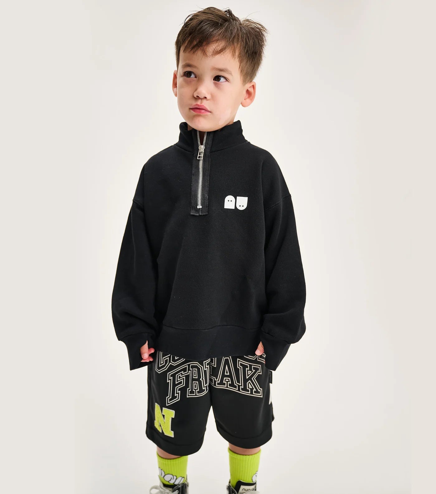 A child wearing a black half zip sweatshirt with a rounded collar, thumbholes, and graphic details on the lower part. The sweatshirt is in a gender-neutral cut.