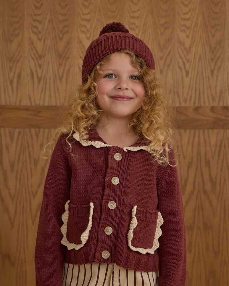 Young girl wearing a maroon knitted cardigan and hat against a wooden paneled wall.