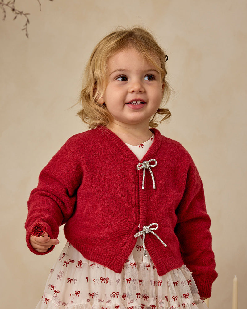 Child wearing a red knitted cardigan with bow details against a beige background