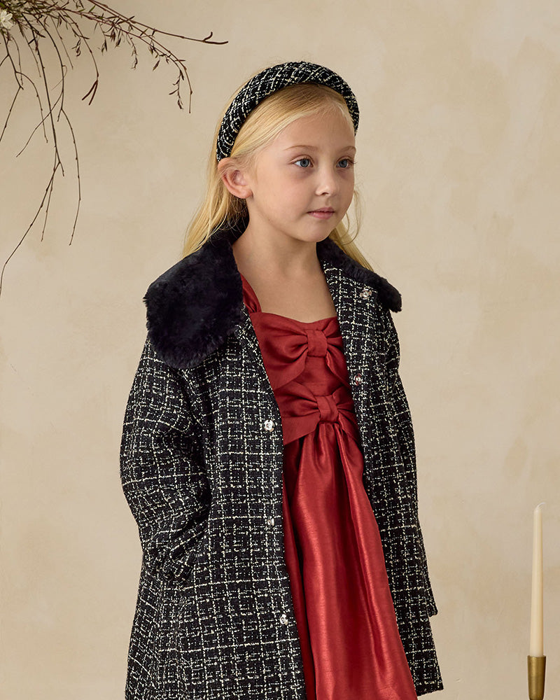 Young girl wearing a black coat with fur collar and a red dress against a beige background