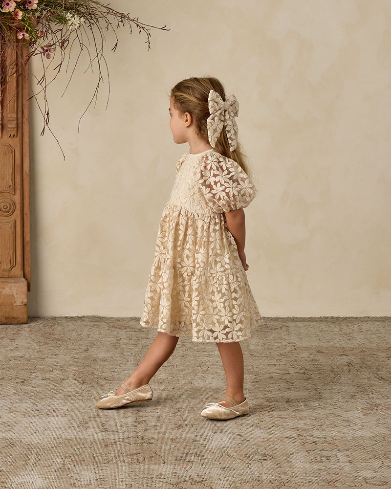 Young girl in a floral dress standing against a beige wall with a rustic wooden cabinet.