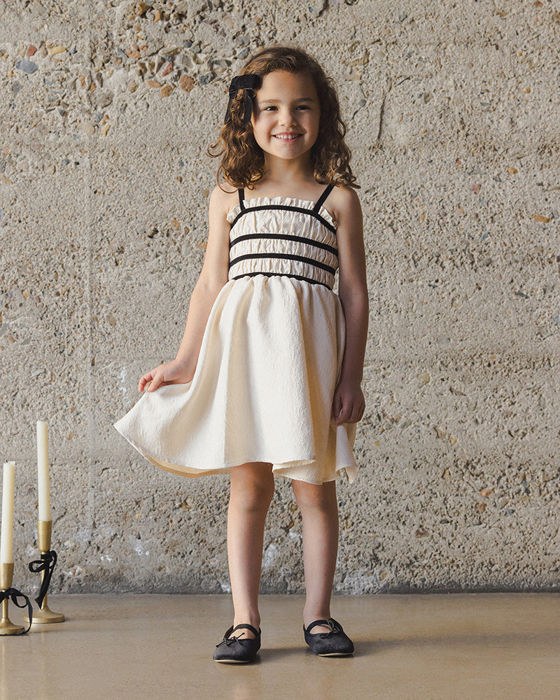 Young girl in a white dress with black straps standing against a textured wall.