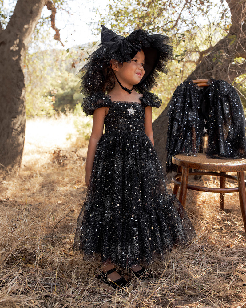 A young girl stands outdoors in a wooded setting wearing a black off-the-shoulder tulle dress covered in silver star-like embellishments, with a large matching black bow headpiece.
