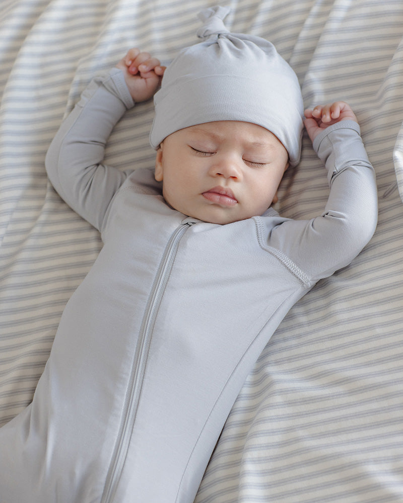 A sleeping newborn baby wearing a light gray long-sleeve zip-up sleeper and matching knotted beanie, lying on a beige and white striped blanket.
