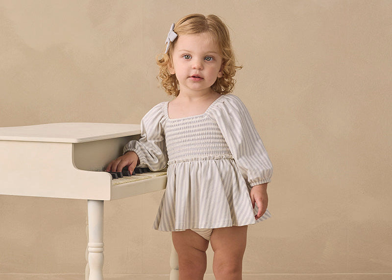 A toddler girl with blonde curly hair stands next to a small white piano in a studio setting. She is wearing a cream and white vertically striped smocked long-sleeve romper dress, cream socks, and tan shoes.