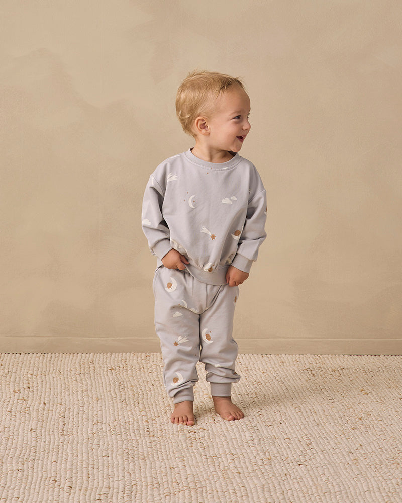 Light gray baby jogger pants with a celestial print of crescent moons, stars, and clouds, featuring an elastic waistband with drawstrings and ribbed ankle cuffs.