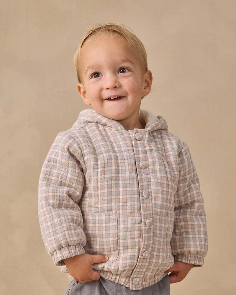 A smiling toddler wearing a beige and brown plaid hooded button-up jacket, standing against a neutral beige studio background.