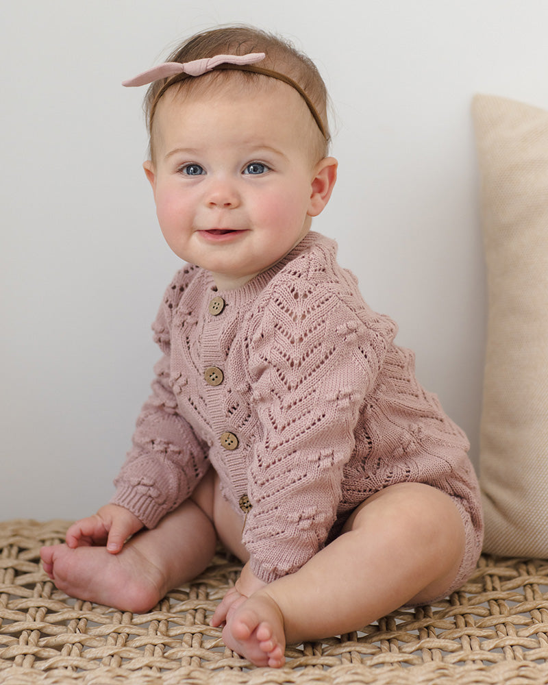 Baby wearing a pink knitted cardigan sitting on a textured surface with a neutral background