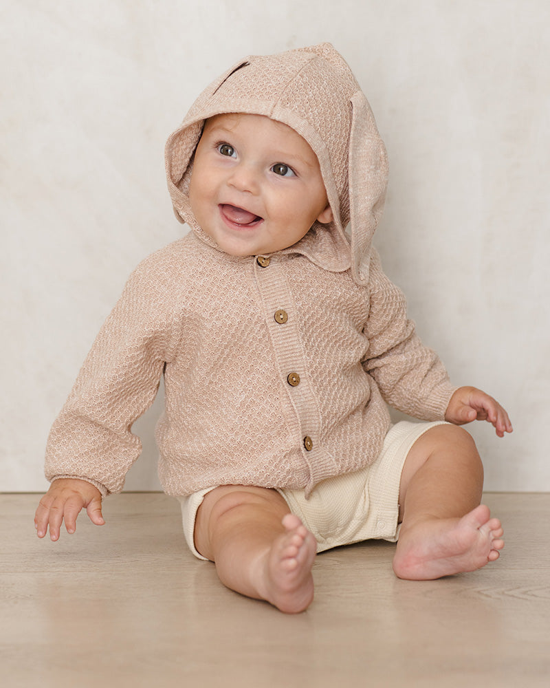 Baby wearing a beige knitted hoodie with bunny ears on a light wooden floor.