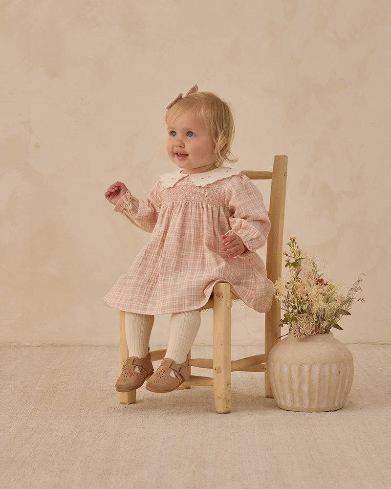 Young child in a pink checkered dress sitting on a wooden chair with a vase of flowers on a beige background
