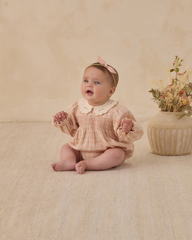 Baby in a pink checkered outfit with a white collar sitting on a beige floor next to a vase of flowers.