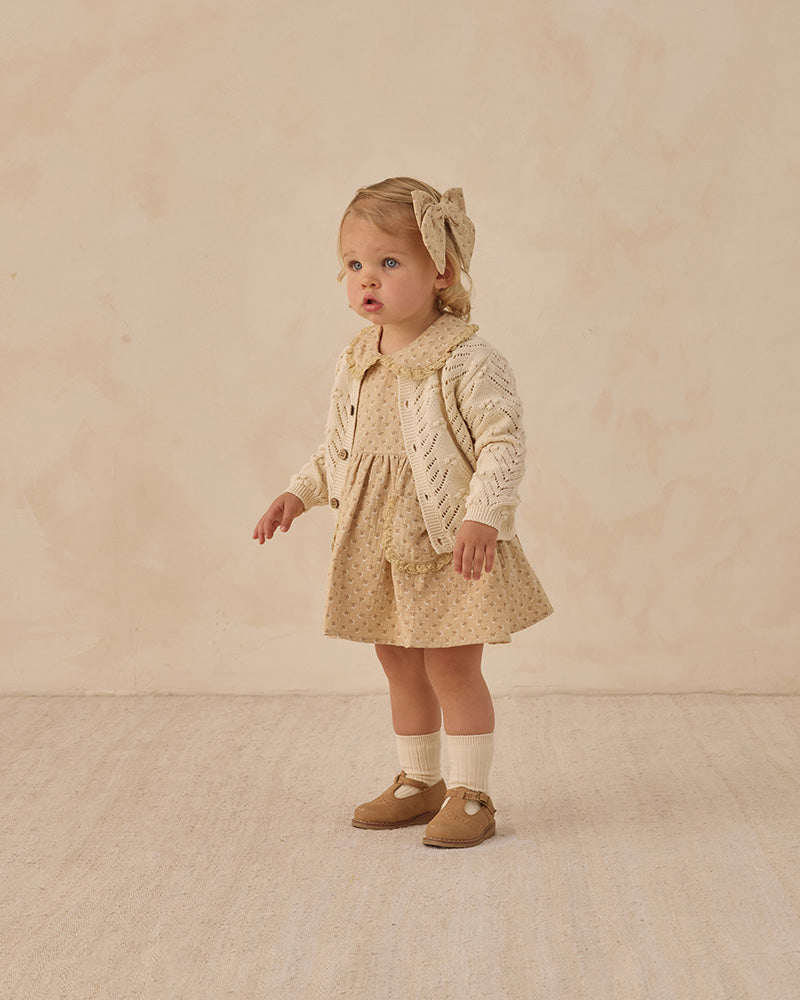Young child wearing a beige dress with a patterned cardigan against a plain background