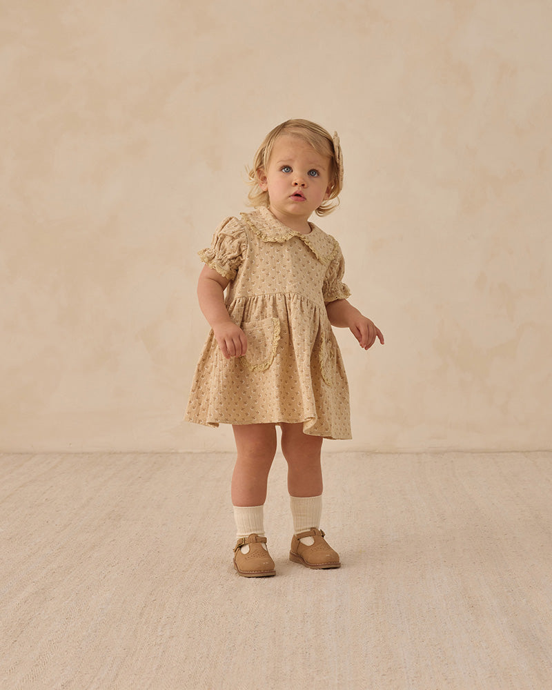 Young child wearing a beige dress with ruffled collar against a beige background