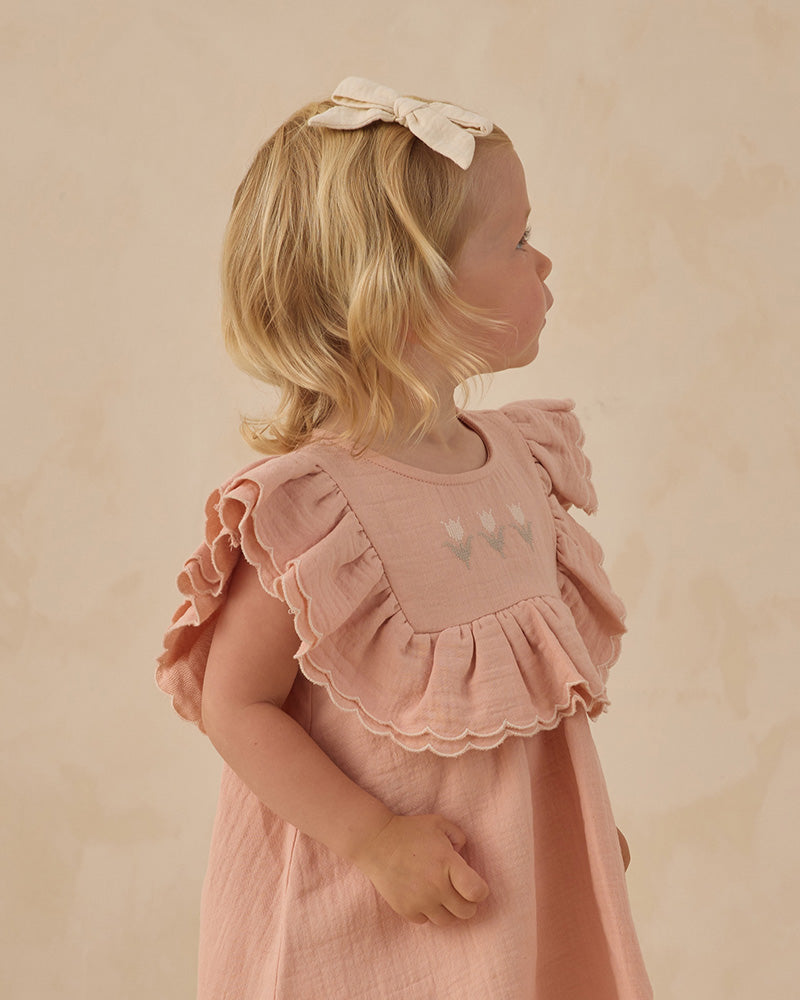 Young girl wearing a pink dress with ruffled sleeves and a bow in her hair against a beige background