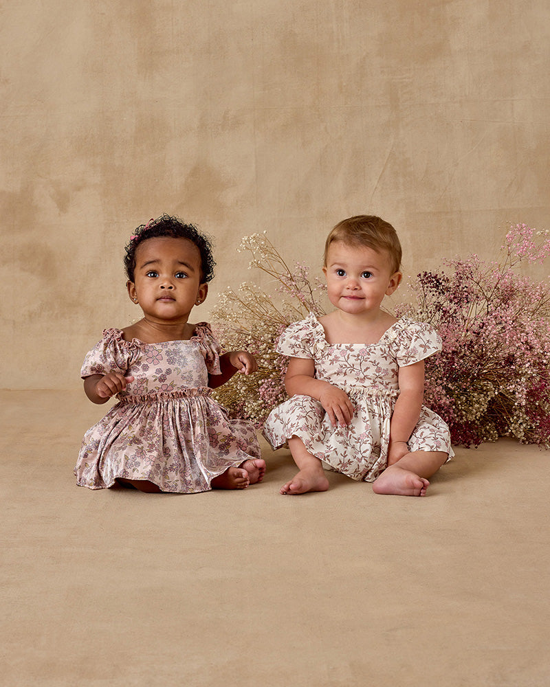 Two young children in floral dresses sitting on a beige surface with flowers in the background.