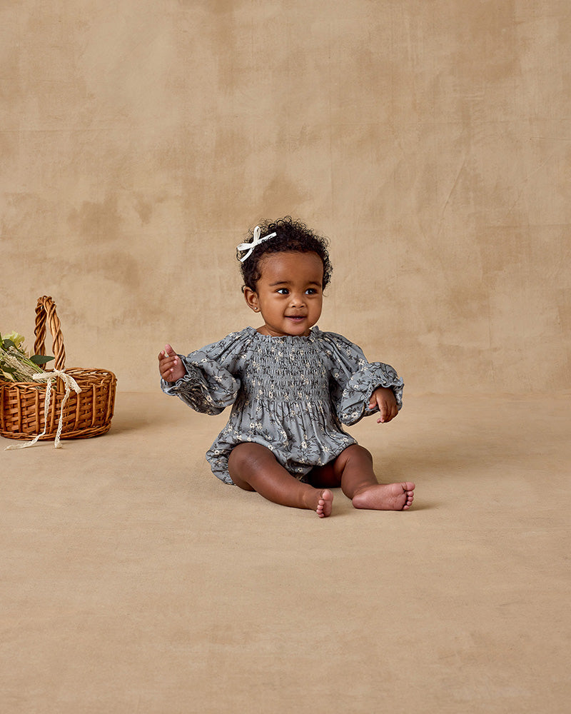 Baby in a floral outfit sitting on a beige background with a woven basket and flowers.