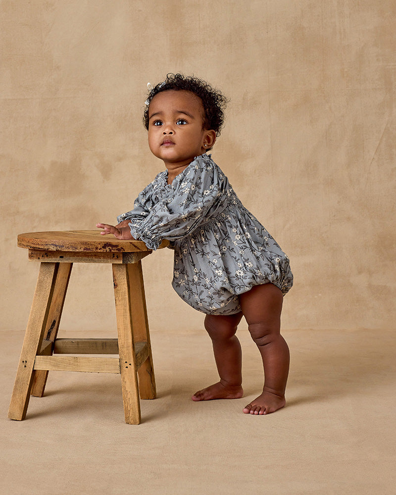 Baby in a floral romper standing next to a wooden stool on a beige background