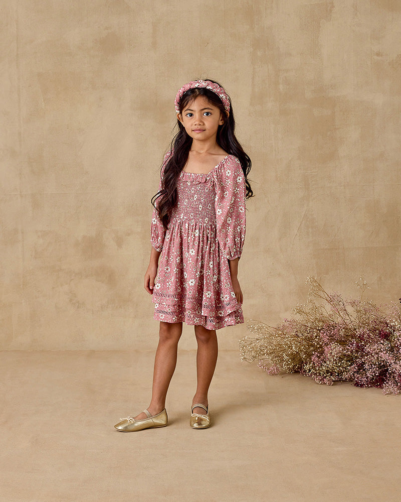 Young girl in a pink floral dress standing against a beige background with dried flowers.