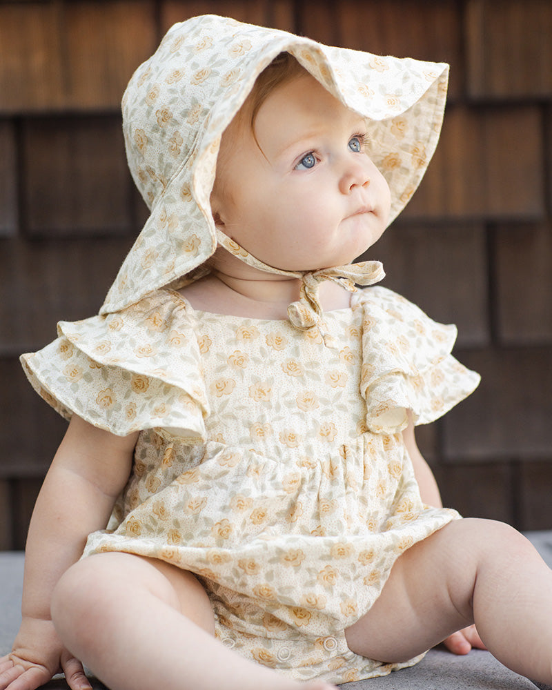 Baby wearing a floral sun hat and romper with ruffle sleeves sitting against a wooden wall.