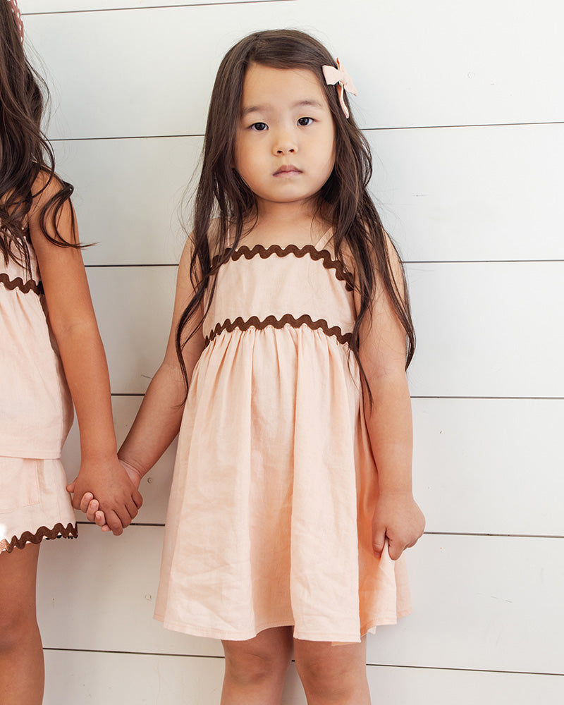 A young girl wearing matching peach dresses with trim against a white wooden background.