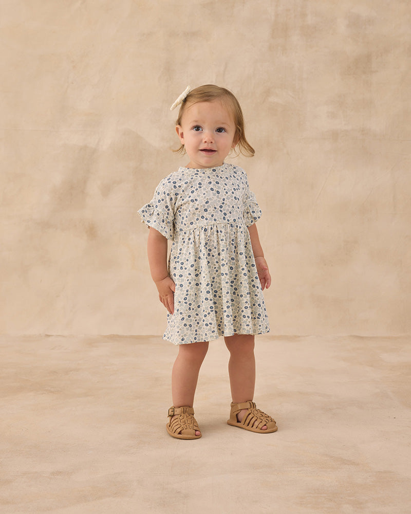 Young child wearing a blue and beige floral dress against a beige background
