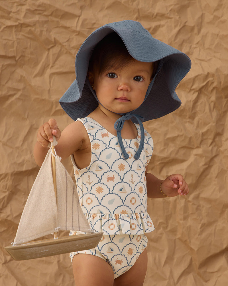 Child wearing a floral swimsuit and blue sun hat holding a toy sailboat against a brown textured background