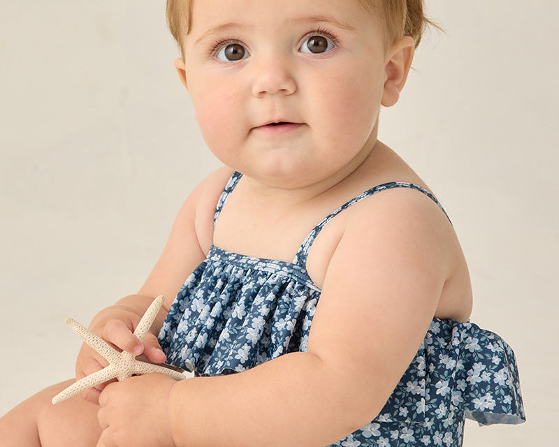 Baby wearing a blue floral dress with a bow on a neutral background
