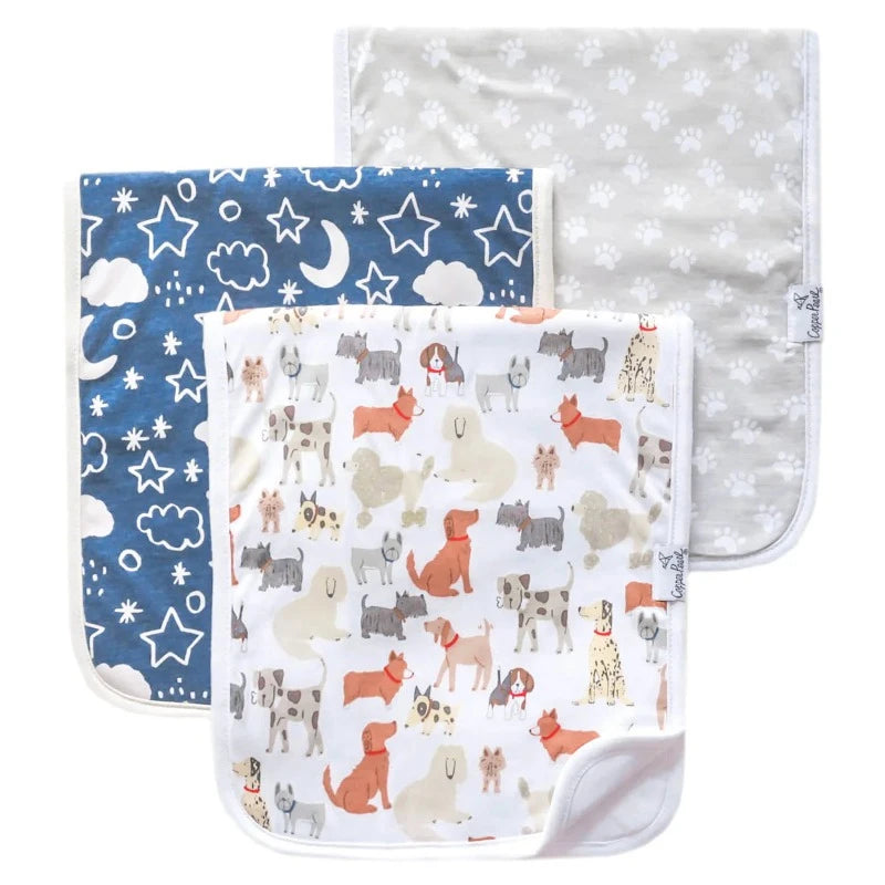 A set of three burp cloths with different patterns. One has a navy blue pattern with stars and moons, another with a beige animal pattern, and the last with a grey paw pattern.