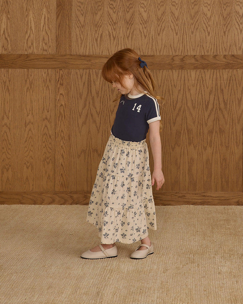Young girl in a floral skirt and navy top standing against a wooden paneled wall.