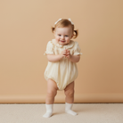 Baby in a beige detailed romper standing against a beige wall