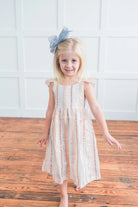 Young girl in a striped dress with a blue bow standing on a wooden floor against a white wall.