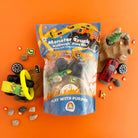 The image shows the Monster Truck KidDough Play Kit contents including a bag of textured brown play dough, monster truck, tires, tree, road signs, drive-through ring of fire, jar of fire fimos, crash car, and natural rocks, all arranged on an orange background.